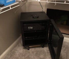 Black AV equipment cabinet with glass door open, placed in a carpeted closet corner under wire shelving.