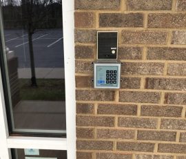 Keypad entry system and intercom mounted on a brick wall next to a door with a safety reminder sign for parents.