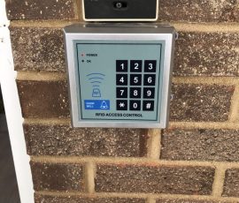 RFID access control keypad and intercom mounted on a brick wall.
