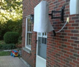 A wall-mounted TV, still covered in foam packaging, is attached to an exterior brick wall near a backyard door.