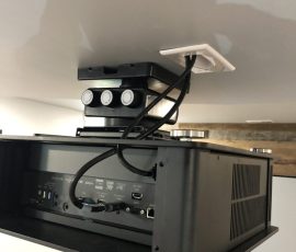 Ceiling-mounted projector with cables connected to a nearby wall outlet on a white ceiling.