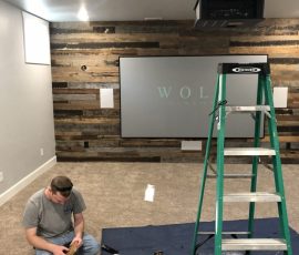 A man assembles equipment on the floor of a home theater room with a ladder, projector, and screen in view.
