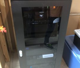 A black server rack cabinet with glass door, housing network equipment, is placed in a room with wooden walls.