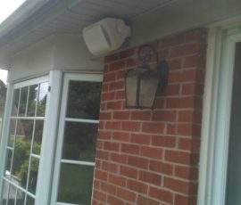 A white outdoor speaker is mounted under an eave near a window and a wall-mounted lantern on a brick house.