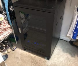 A server rack cabinet with blue LED lights is placed on the floor in a walk-in closet surrounded by clothes and shoes.