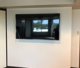 Flat screen TV mounted on a plain white wall in an office with carpeted floor; papers lying on the floor below.