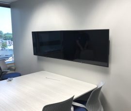 Conference room with a white table, blue chairs, and two wall-mounted flat screen monitors.