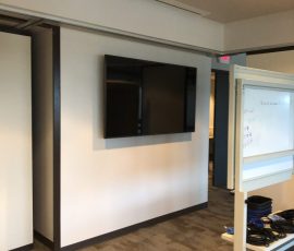 A wall-mounted flat screen TV is displayed in an office space with carpeted floors and a mobile whiteboard nearby.