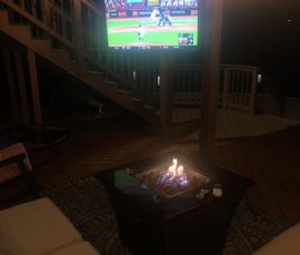Outdoor patio with a fire pit, couch, TV showing a baseball game, and wooden stairs in the background.