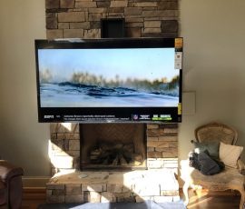Flat-screen TV mounted above a stone fireplace, with tools and a blanket on the floor in a sunlit living room.
