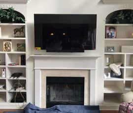 A flat-screen TV is mounted above a fireplace between built-in shelves with books, photos, and plants. Tools are on the floor.