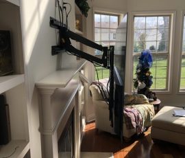 Flat-screen TV mounted on an adjustable wall arm above a fireplace in a sunlit living room with large windows.