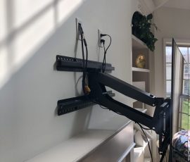 Wall-mounted TV on an adjustable arm with visible cables, next to a shelf and a window in a sunlit room.
