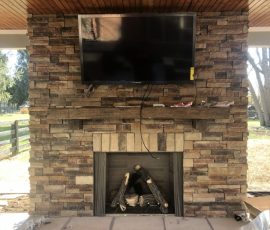 Stone fireplace with a wall-mounted TV above the mantel, under a wooden ceiling with heaters, in an outdoor space.