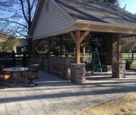 Outdoor covered patio with stone pillars, bar area, wall-mounted TV, and nearby patio seating on a sunny day.
