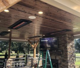 A green ladder stands under a wooden ceiling with heaters and lights, next to a stone fireplace in an outdoor patio area.