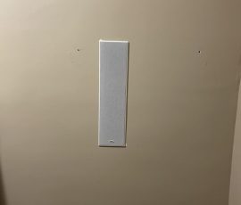 A rectangular white wall-mounted speaker is centered on a plain beige wall with two small holes on either side.