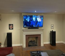 A living room with a TV mounted above a fireplace, displaying three people in a scene, and electronics on a coffee table.