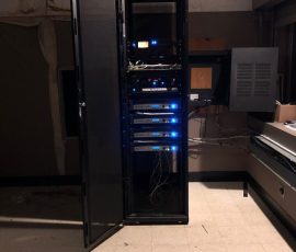 A server rack with several network devices and cables stands in a dimly lit, sparsely furnished room with tile flooring.