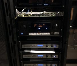 A server rack with network equipment, audio receiver, and multiple rack-mounted power amplifiers with indicator lights.