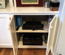 Open cabinet showing electronic devices, remote, manuals, and miscellaneous items on two shelves below a framed horse painting.
