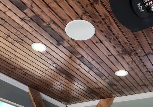 Wood-paneled outdoor ceiling with recessed lights, a round speaker, wood beams, and rooftops visible outside.