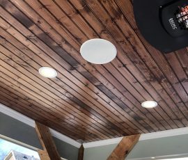 Wood-paneled outdoor ceiling with recessed lights, a round speaker, wood beams, and rooftops visible outside.