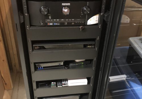 Black server rack with multiple electronic components, control knobs, and remote controls in a room with wooden walls.