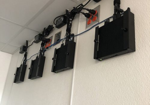 Four black electronic devices are mounted on a white wall, each connected to power outlets and network cables above.