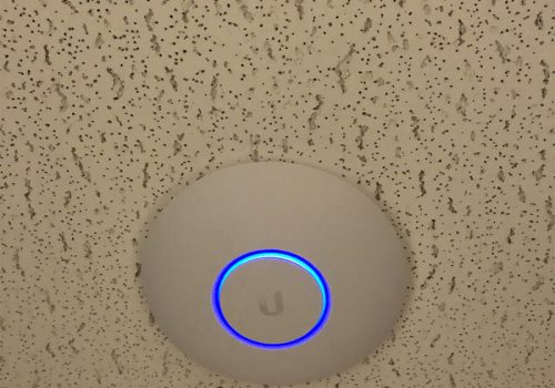 A round wireless access point with a blue LED ring is mounted on a speckled ceiling.
