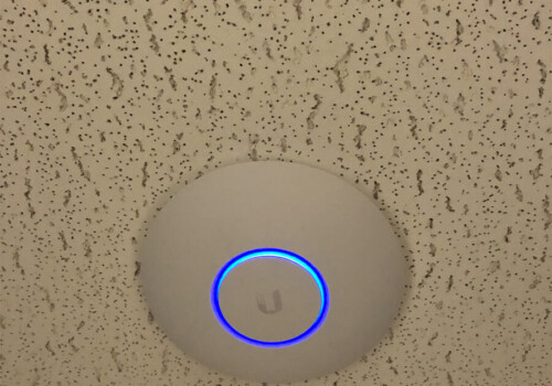 A round wireless access point with a blue LED ring is mounted on a speckled ceiling.