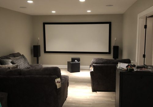 Home theater room with a large screen, two couches, speakers, and a small table, with equipment on the floor in front.