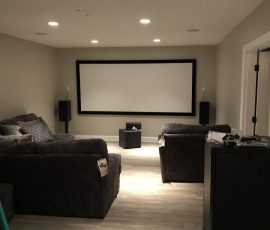 Home theater room with a large screen, two couches, speakers, and a small table, with equipment on the floor in front.